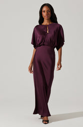 A woman is wearing a deep purple satin dress with a loose-fitting, draped top that has wide sleeves and a keyhole neckline, paired with a fitted, high-waisted, long skirt that reaches the floor. She is also wearing black open-toe heels.